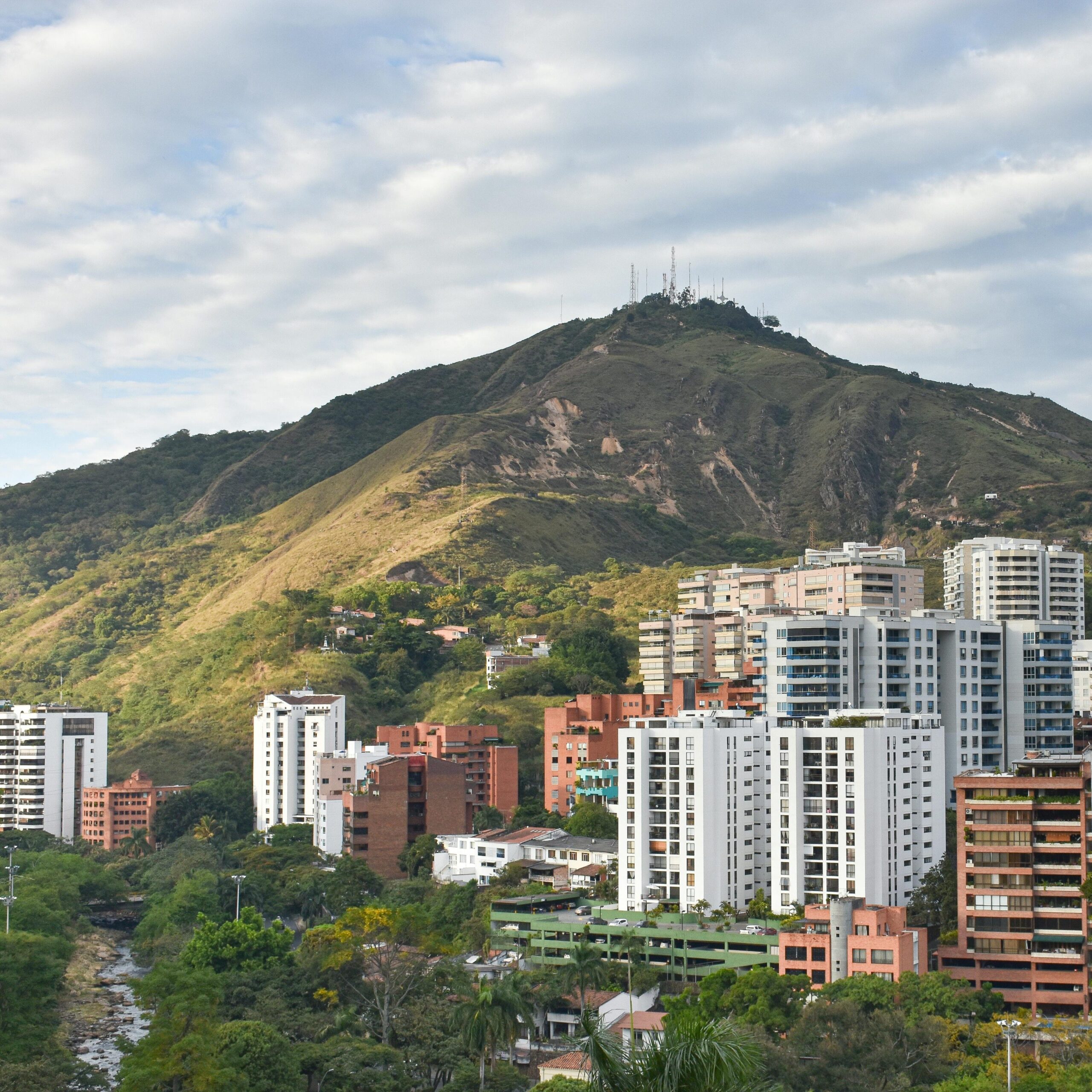 A vibrant cityscape of Cali, Colombia, with lush Andes mountains and modern buildings under a cloudy sky.